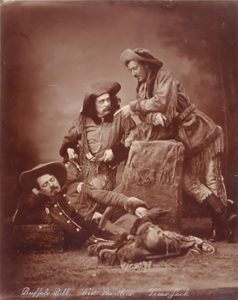 Three men are in a group in front of the camera wearing 1800s style cowboy clothes. One man is standing while leaning forward on a box. One man is sitting and leaning on the box. One man is lying on his side, head propped on his hand.