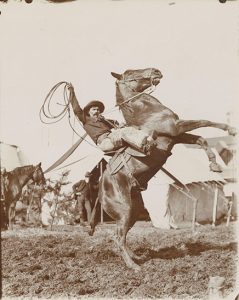 A man sitting in a saddle on a horse, holding a looped lasso over his head. The horse is rearing up on his back legs.