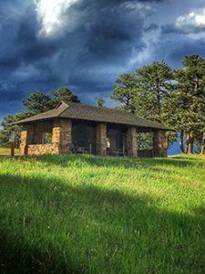 Open air structure made of large stones sitting in a grassy field. Trees behind it with a dark cloudy sky.