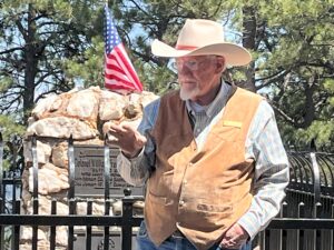 A man standing in from of a black fence. He is wearing a long sleeve shirt, vest, and cowboy hat. Beyond the fence is a stone grave marker with an American flag on top of it.
