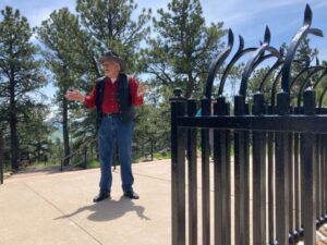 Man standing next to a large black fence. He is wearing jeans, a red button up shirt, vest, and a cowboy hat.
