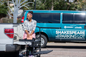 A woman is kneeling on the tailgate of a white truck. Blue van in background with Shakespeare in the Parking Lot written in white letters.